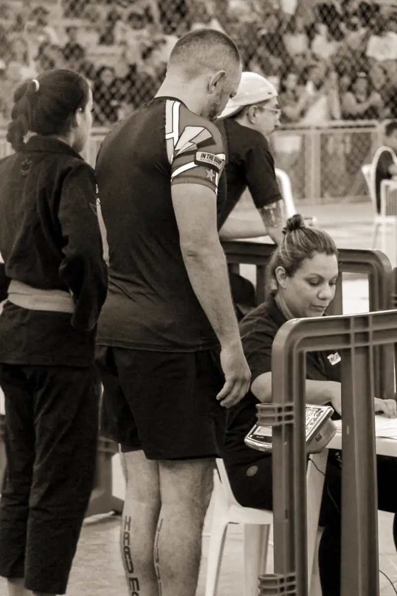 Pécora de rash guard e shorts junto a uma mesa no ginásio da competição, com a arquibancada ao fundo